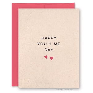 May include: A greeting card with a pink envelope. The card has the words "HAPPY YOU + ME DAY" in black text, with two small pink hearts below. The card is a beige color.