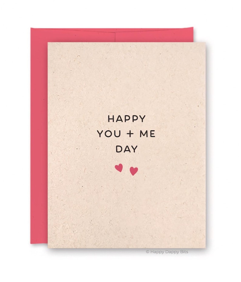 May include: A greeting card with a pink envelope. The card has the words "HAPPY YOU + ME DAY" in black text, with two small pink hearts below. The card is a beige color.