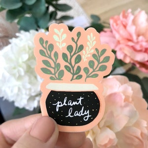May include: A peach-coloured sticker with a black pot and green plant. The pot has white speckles and the text "plant lady" in white.