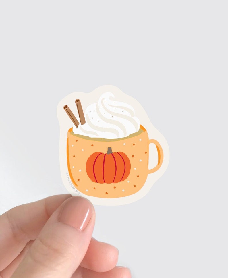 May include: A white ceramic mug with a pumpkin design and a brown handle. The mug is filled with a white whipped cream topping and two cinnamon sticks.
