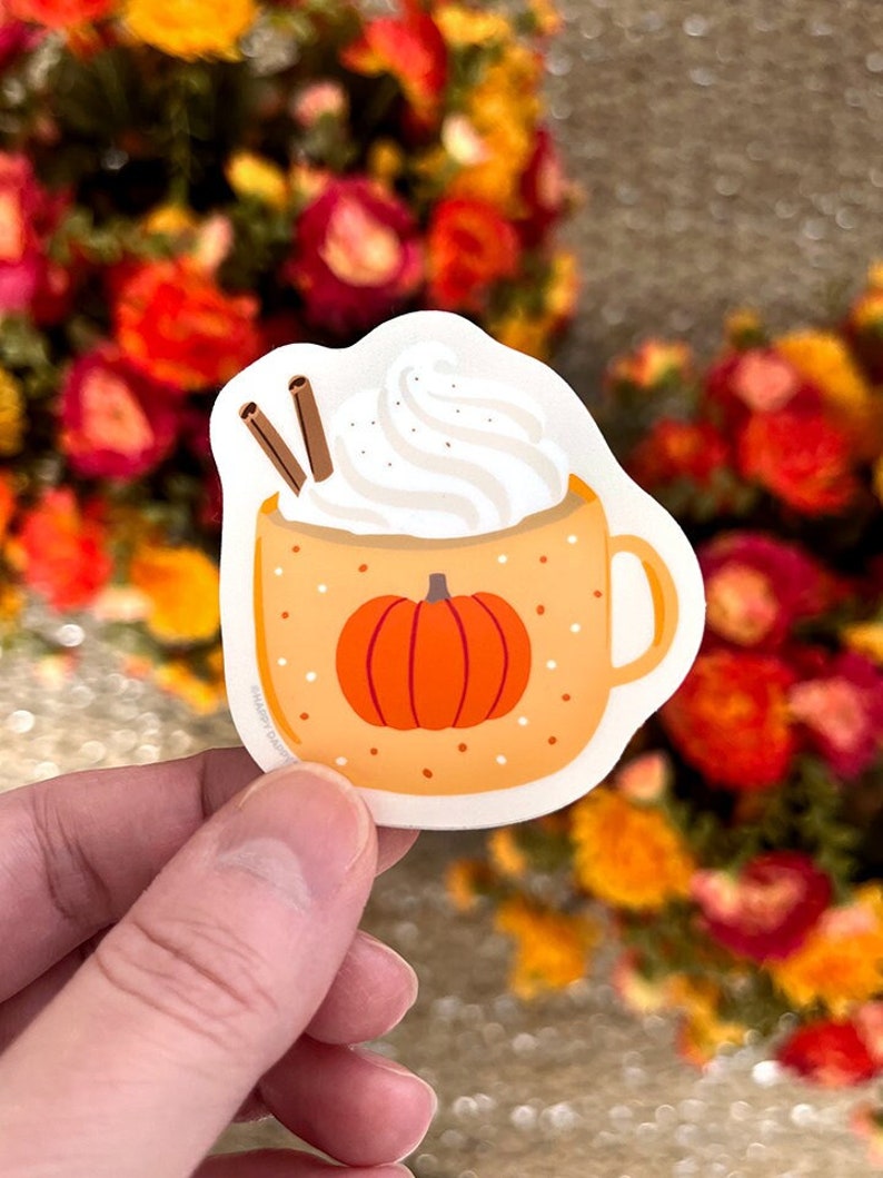May include: A sticker of a pumpkin spice latte with whipped cream and cinnamon sticks. The mug is orange with white polka dots and a pumpkin on the front.