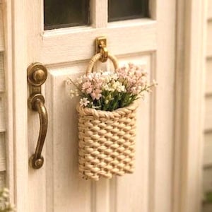 Könnte beinhalten: Eine weiße Miniatur-Hausfassade mit einer Tür mit vier Fensterscheiben, einem Messinggriff und einem hängenden geflochtenen Korb voller kleiner rosa und weißer Blumen. Eine schwarze Wandlampe, ein Schild mit der Nummer 24, zwei Pflanzen und eine Fußmatte.