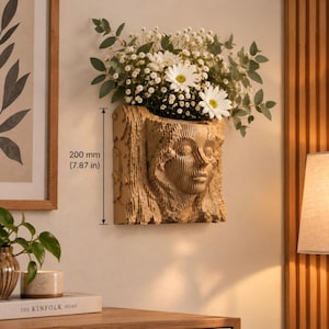 Puede incluir: Una maceta de pared beige con un rostro humano esculpido integrado en un diseño de corteza de árbol texturizada. La maceta mide 200 mm de altura y contiene un ramo de margaritas blancas y hojas de eucalipto verdes.