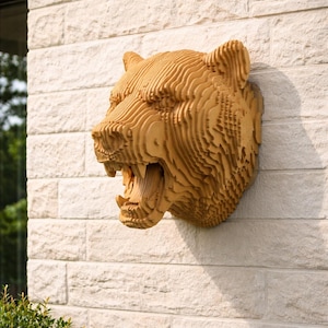 Pode incluir: Uma escultura de cabeça de urso montada na parede, construída com camadas empilhadas de cor castanho-claro cortadas a laser. O urso é representado com a boca aberta, revelando os dentes, e o design em camadas cria uma textura topográfica.