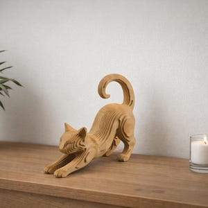 May include: A wooden cat figurine in a stretching pose. The light brown cat sculpture is on a wooden surface, with a houseplant in a white pot and a lit candle in a glass nearby. The cat's tail is curled.