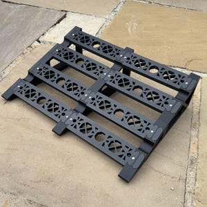 May include: A black metal pallet-style decorative piece with an openwork design. The pallet has a series of horizontal bars with geometric cutouts and is supported by short legs. The design includes small, decorative rivets.