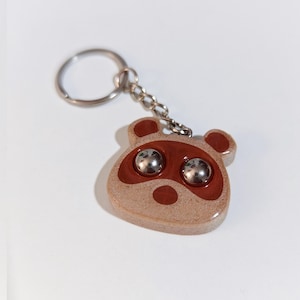Puede incluir: Un llavero marrón y beige con un diseño de cara de mapache, con dos ojos de bola de metal plateado. El llavero tiene un anillo y una cadena de metal plateado.