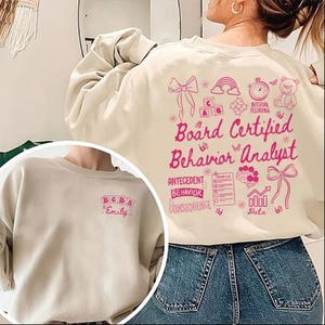 May include: A beige sweatshirt with pink text and graphics. The back of the sweatshirt features the words "Board Certified Behavior Analyst" with related illustrations. The front has "BCBA" and a name. The sweatshirt is made of a soft material.