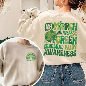 Op de afbeelding: Crèmekleurige sweatshirt met groene tekst en afbeeldingen die Cerebral Palsy Awareness promoten. De achterkant toont de zin "In March We Wear Green Cerebral Palsy Awareness". De voorkant heeft een kleiner ontwerp met een regenboog en de woorden "Cerebral Palsy Awareness" en "Team Alex."