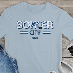 May include: Light blue t-shirt with the text "SOKCER CITY USA" in a distressed, two-tone design. The shirt has a crew neck and short sleeves. The graphic is in white and blue.