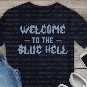 Puede incluir: Camiseta azul marino con el texto "WELCOME TO THE BLUE HELL" en azul claro con letras de estilo llama. La camiseta tiene cuello en V. Una prenda casual con un diseño llamativo.