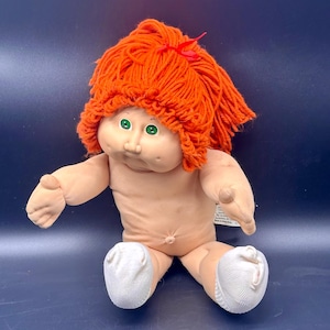 Può includere: Bambola vintage Cabbage Patch Kid con capelli di filo arancione e occhi verdi. La bambola ha un corpo morbido color carne e indossa calzini bianchi. Un piccolo fiocco rosso adorna la parte superiore dei capelli della bambola.