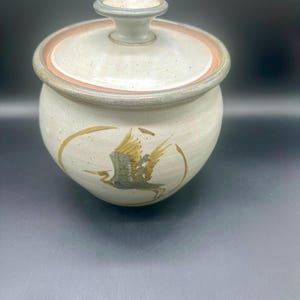 May include: A ceramic jar with a lid. The jar is off-white with a brown rim and a handle on the lid. A bird is painted on the side in shades of brown and green. The jar is round and has a speckled texture.