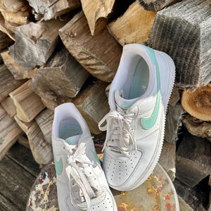 May include: A pair of white sneakers with mint green accents, including the iconic Nike swoosh. The shoes are laced up and sit on a weathered metal surface, with a backdrop of stacked firewood.