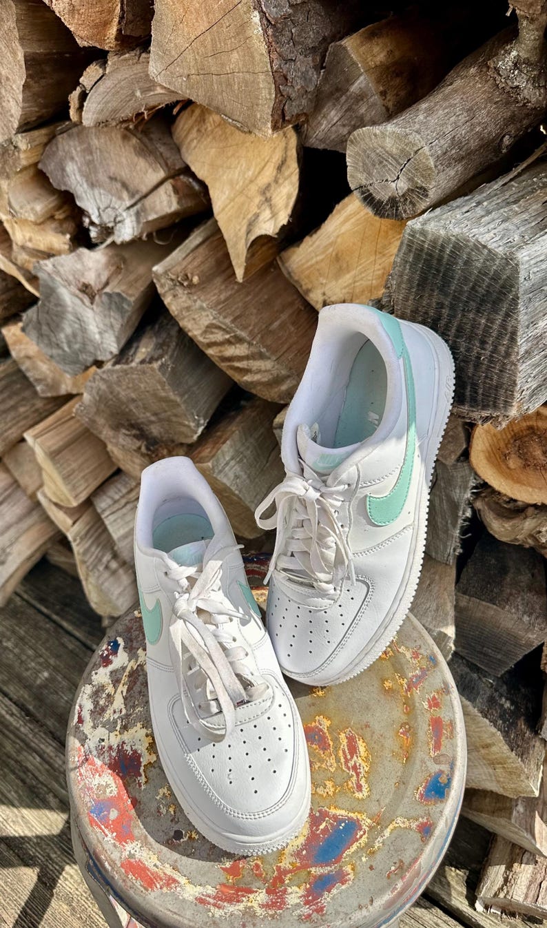 May include: A pair of white sneakers with mint green accents, including the iconic Nike swoosh. The shoes are laced up and sit on a weathered metal surface, with a backdrop of stacked firewood.