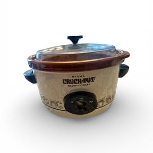 Op de afbeelding: Een Rival Crock-Pot slowcooker met een bruine rand en een doorzichtig glazen deksel. De crèmekleurige basis heeft een decoratief patroon en zwarte handgrepen. De bedieningsknop heeft instellingen voor Laag, Uit en Hoog.