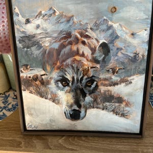 May include: A framed painting of a wolf in a snowy mountain landscape. The wolf's face is in the foreground, with detailed fur and intense eyes. The background features snow-covered mountains and a cloudy sky. The artwork is signed in the lower left corner.