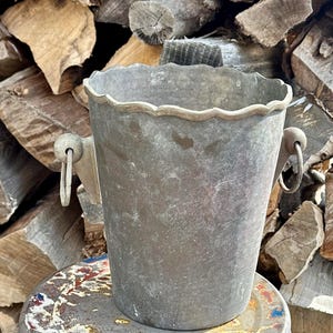 Puede incluir: Un cubo de metal desgastado con un borde festoneado y dos asas con anillas. El cubo tiene forma cilíndrica y un aspecto envejecido y desgastado. El fondo presenta una pila de leña.