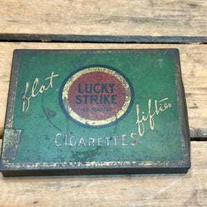 May include: Vintage Lucky Strike cigarette tin from the 1950s. The rectangular tin has a green background with gold lettering and a red and gold circular logo. The text on the tin reads "flat fifties" and "Cigarettes".