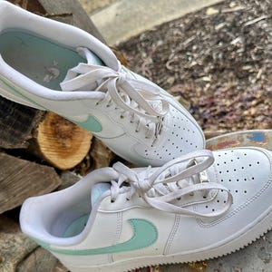 May include: A pair of white sneakers with light blue accents. The shoes have a classic design with a perforated toe box, white laces, and a light blue swoosh logo on the side. The soles are white and the insoles are light blue.