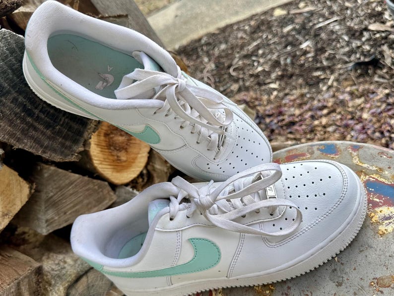 May include: A pair of white sneakers with light blue accents. The shoes have a classic design with a perforated toe box, white laces, and a light blue swoosh logo on the side. The soles are white and the insoles are light blue.