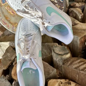 May include: Pair of white sneakers with light blue accents, including the iconic swoosh logo. The shoes feature a classic design with laces and are set against a natural wood background. A stylish and comfortable footwear option.