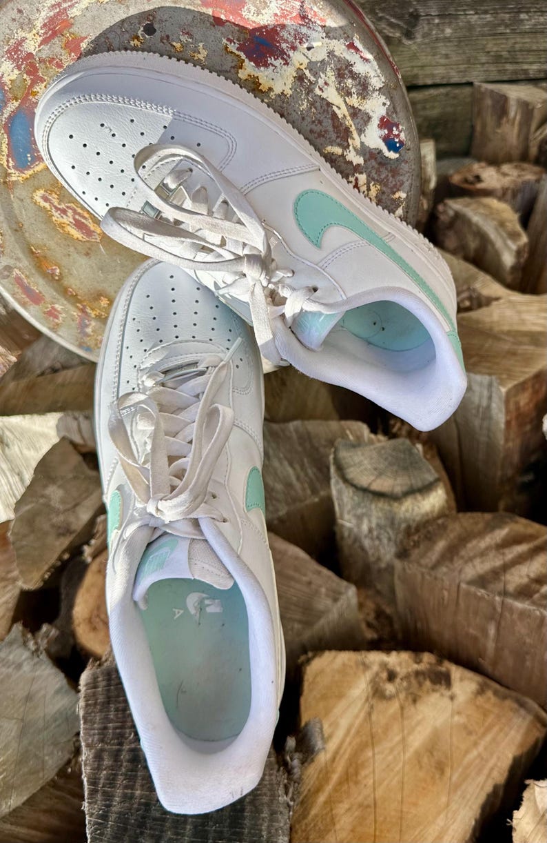 May include: Pair of white sneakers with light blue accents, including the iconic swoosh logo. The shoes feature a classic design with laces and are set against a natural wood background. A stylish and comfortable footwear option.