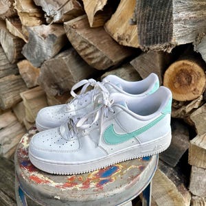 May include: White leather sneakers with a mint green swoosh and heel detail. The shoes have white laces and are sitting on a small, round, distressed metal stool. The background is a stack of firewood.