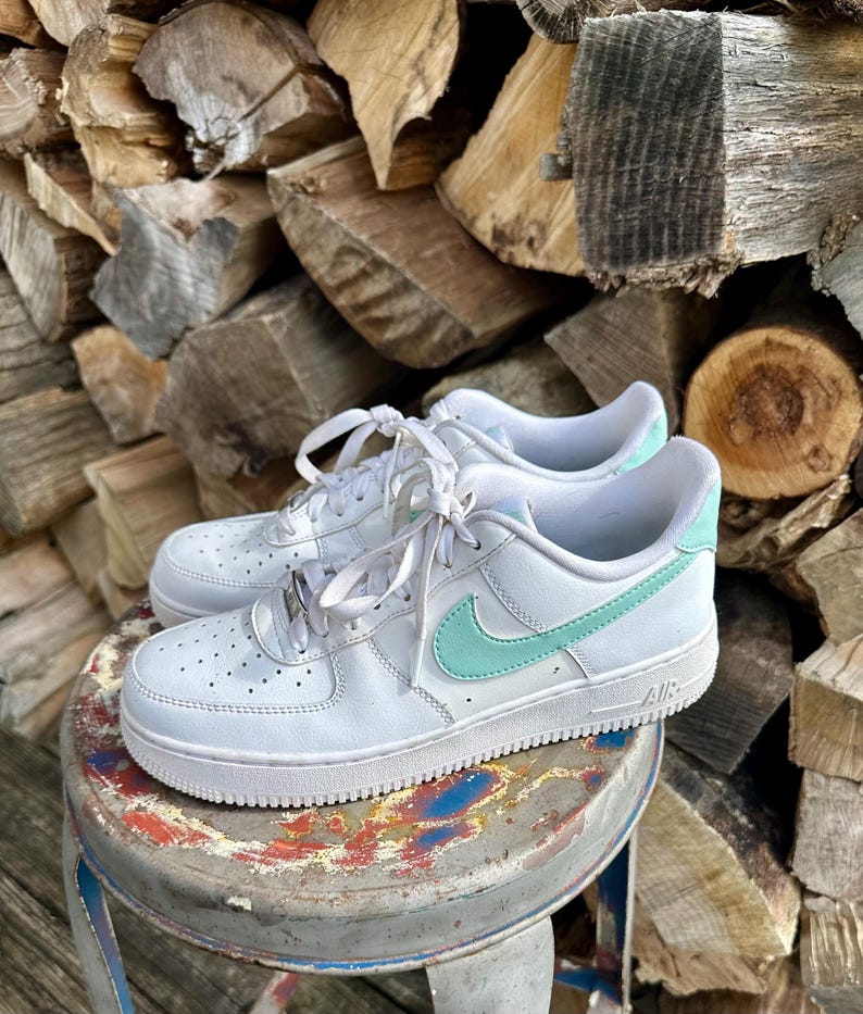 May include: White leather sneakers with a mint green swoosh and heel detail. The shoes have white laces and are sitting on a small, round, distressed metal stool. The background is a stack of firewood.