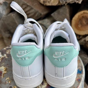 May include: A pair of white Nike Air Force 1 sneakers. The heel tab is a light teal color with the Nike Air logo in white. The laces are white, and the shoes are set against a background of stacked wood.