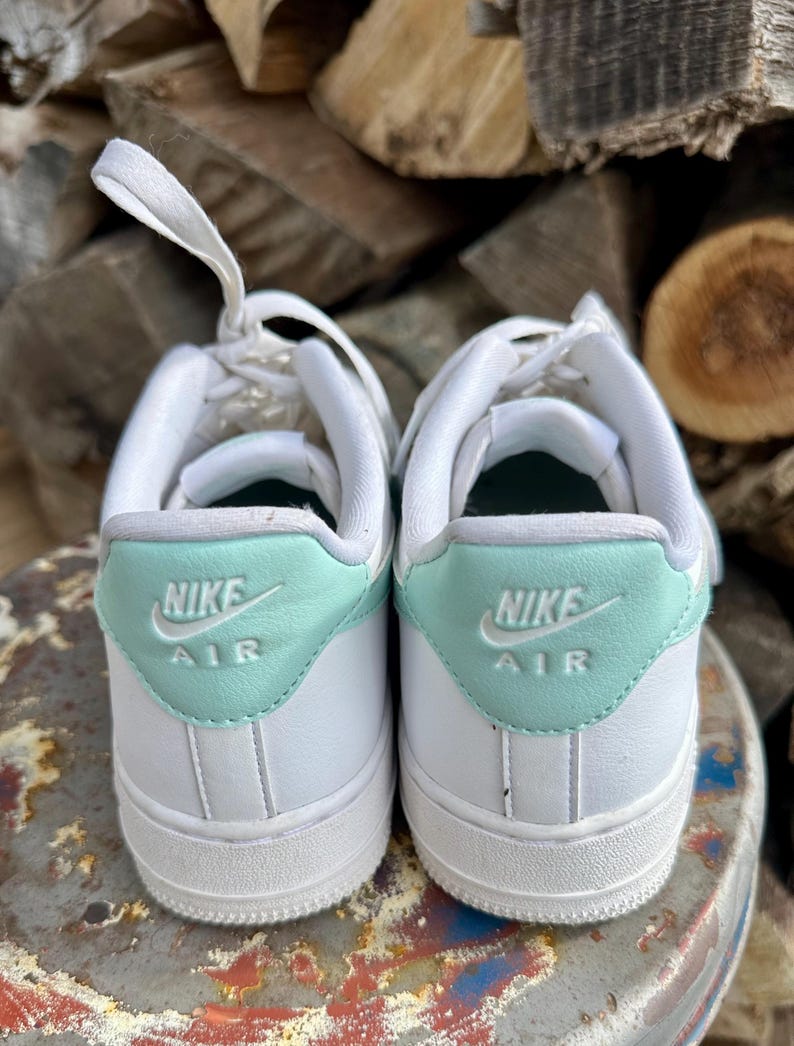 May include: A pair of white Nike Air Force 1 sneakers. The heel tab is a light teal color with the Nike Air logo in white. The laces are white, and the shoes are set against a background of stacked wood.