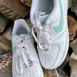 May include: Pair of white leather sneakers with light blue accents, including the iconic Nike swoosh logo. The shoes have white laces and soles, and are placed on a metal surface with a wood background.