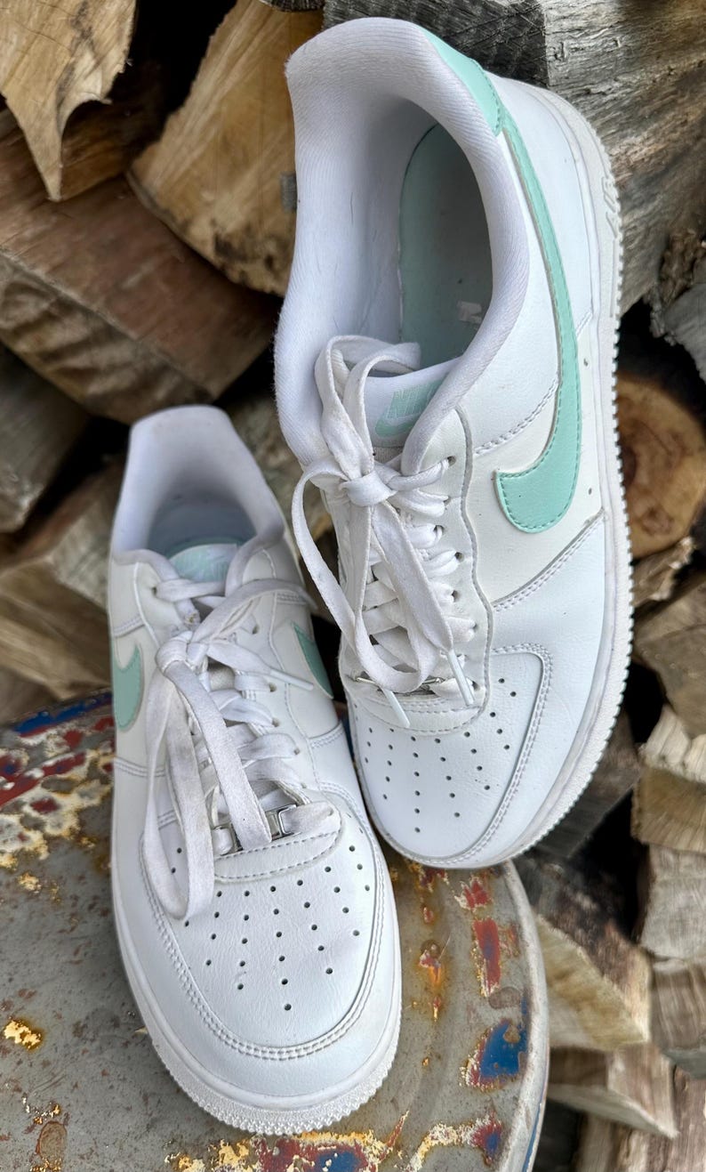 May include: Pair of white leather sneakers with light blue accents, including the iconic Nike swoosh logo. The shoes have white laces and soles, and are placed on a metal surface with a wood background.