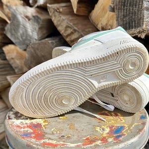 May include: White Nike Air Force 1 sneakers with mint green accents. The sneakers are placed on a small, round, distressed stool. The soles of the shoes are visible, showing the Nike logo and tread pattern.