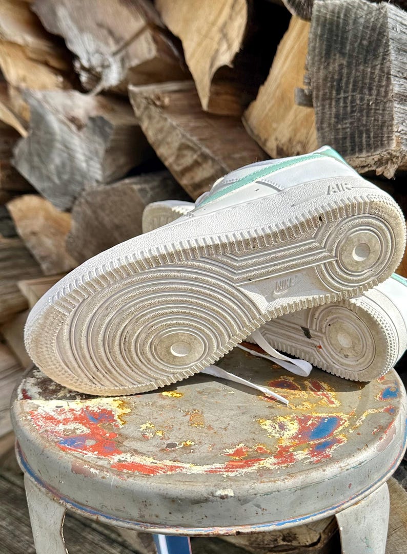 May include: White Nike Air Force 1 sneakers with mint green accents. The sneakers are placed on a small, round, distressed stool. The soles of the shoes are visible, showing the Nike logo and tread pattern.