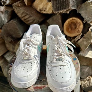 May include: Pair of white leather sneakers with light blue accents on the side and tongue. The sneakers have white laces and a perforated design on the toe box. The shoes are sitting on a metal surface.