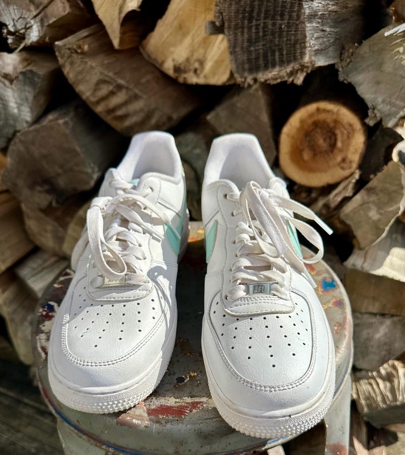 May include: Pair of white leather sneakers with light blue accents on the side and tongue. The sneakers have white laces and a perforated design on the toe box. The shoes are sitting on a metal surface.