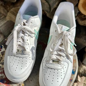 May include: Pair of white leather sneakers with mint green accents, including the Nike swoosh logo. The shoes have white laces and a perforated toe design. The sneakers are on a weathered surface.