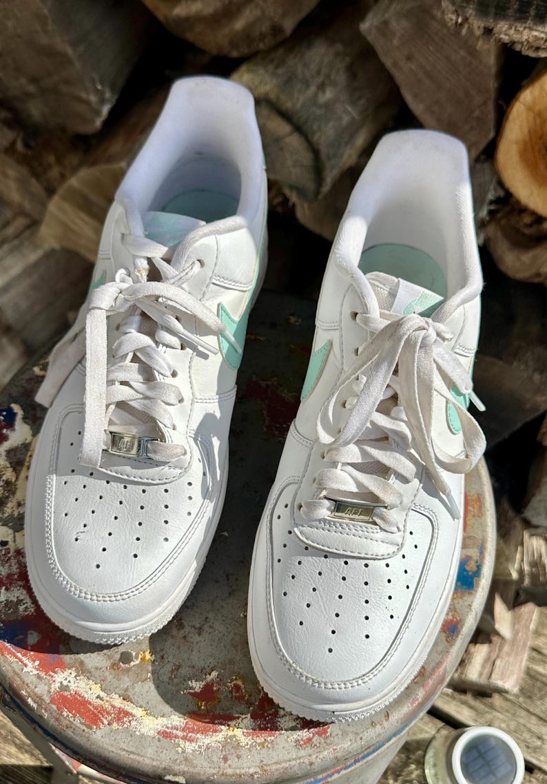 May include: Pair of white leather sneakers with mint green accents, including the Nike swoosh logo. The shoes have white laces and a perforated toe design. The sneakers are on a weathered surface.