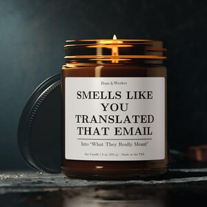 Puede incluir: Una vela de soja encendida en un tarro de cristal marrón con tapa negra. La etiqueta dice "SMELLS LIKE YOU TRANSLATED THAT EMAIL" con el texto "Into 'What They Really Meant'". La vela es de 255 g.