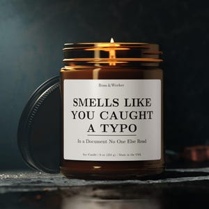 以下が含まれることがあります： 黒い蓋が付いた茶色のガラス瓶に入った、火の灯った大豆キャンドル。ラベルには「SMELLS LIKE YOU CAUGHT A TYPO」と「In a Document No One Else Read」と書かれています。キャンドルは255gです。