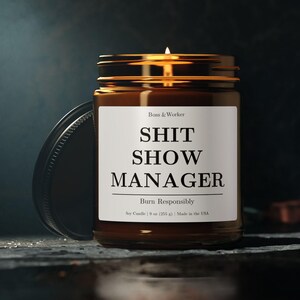 Puede incluir: Una vela de soja encendida en un tarro de vidrio marrón con tapa negra. La etiqueta dice "SHIT SHOW MANAGER" en letras negras en negrita. La vela es de 255 g y está hecha en EE. UU.