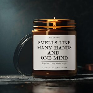 Puede incluir: Una vela de soja encendida en un tarro de cristal marrón con tapa negra. La etiqueta dice "Smells Like Many Hands and One Mind" con el texto "Together They Make Magic". La vela es de 255 g y está hecha en EE. UU.
