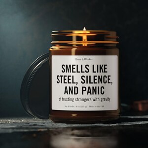 Pode incluir: Uma vela em frasco de vidro castanho com tampa preta e rótulo branco que diz Boss & Worker, SMELLS LIKE STEEL, SILENCE, AND PANIC, of trusting strangers with gravity, Soy Candle | 255 g | Made in the USA, com um pavio aceso.