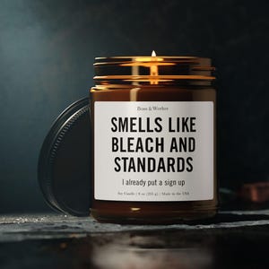May include: A brown glass jar candle with a white label featuring the black text: Boss & Worker, SMELLS LIKE BLEACH AND STANDARDS, I already put a sign up, Soy Candle | 9 oz (255 g) | Made in the USA. The candle has a lit wick and a black lid.