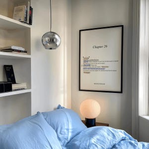 May include: A bedroom scene featuring a light blue bedspread and pillows. A framed print with the text "Chapter 26" hangs on the wall. A spherical lamp sits on a nightstand, and a chrome pendant light hangs from the ceiling.