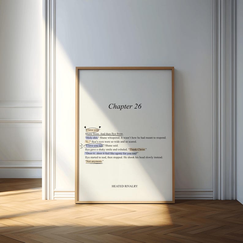 May include: A framed print with a wooden frame, leaning against a white wall. The print features text from "Chapter 26" of a book, including phrases like "I love you" and "Heated Rivalry."