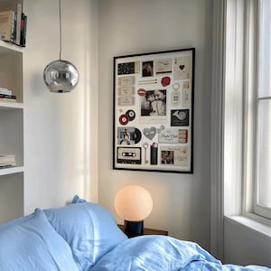 May include: A bedroom scene featuring a large framed collage of photos and memorabilia. A silver orb pendant light hangs from the ceiling. A round, white globe lamp sits on a dark base. The bed is covered with light blue bedding.