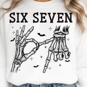 May include: White sweatshirt with the text "SIX SEVEN" in black block letters. The design features a skeleton hand making the "OK" sign with the other hand forming the number seven. Small bats and stars are scattered around the hands.