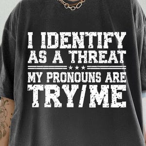 May include: Dark gray t-shirt with white text that reads "I IDENTIFY AS A THREAT. MY PRONOUNS ARE TRY/ME." The text is centered and stacked. The shirt has a crew neck and short sleeves.